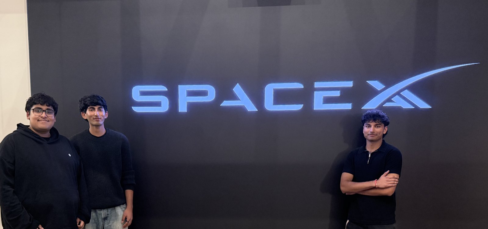 Three young men standing in front of a dark wall with a glowing blue SpaceX logo across it. Two stand together on the left; the third stands alone on the far right with his arms crossed.