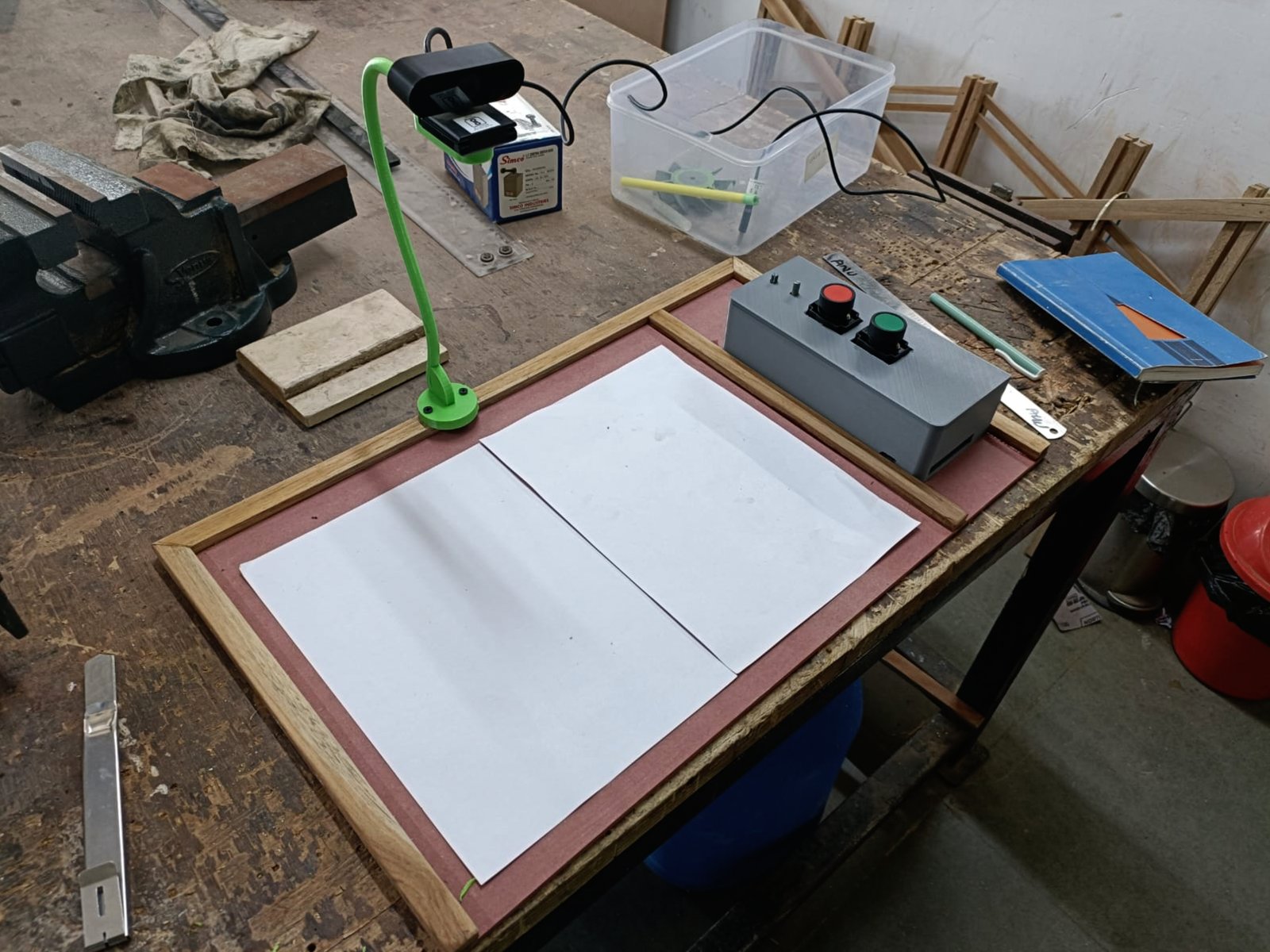 An early TechNetra prototype on a battered workshop bench: a webcam on a bright green gooseneck arm, angled down over a wooden frame holding two blank sheets of paper, next to a grey box with one large red button and one large green button. A metal vise sits at the edge of the bench.